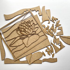 Op de afbeelding: Een houten knutselset met een lotusbloemontwerp. De set bevat meerdere houten onderdelen die kunnen worden geassembleerd om een decoratieve wandhanger of ander knutselproject te maken.