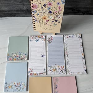 May include: A spiral-bound daily planner with a cream cover featuring a colorful wildflower and butterfly illustration, accompanied by seven assorted rectangular notepads in various pastel colors, each decorated with matching floral and butterfly motifs.