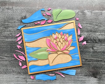 May include: A wooden craft kit featuring a pink and yellow water lily with green leaves, surrounded by blue water. The kit includes separate pieces to assemble the water lily, leaves, and water.