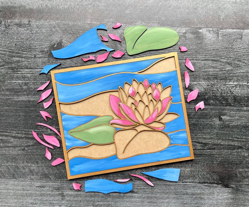 Op de afbeelding: Een houten knutselset met een roze en gele waterlelie met groene bladeren, omringd door blauw water. De set bevat losse onderdelen om de waterlelie, bladeren en het water in elkaar te zetten.
