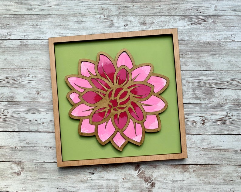 May include: A pink and red wooden flower with layered petals is mounted on a green background in a wooden frame.