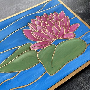 Op de afbeelding: Een handgeschilderd houten paneel met een roze waterlelie en groene bladeren op een blauwe achtergrond met gouden accenten. Het paneel heeft een gouden rand.