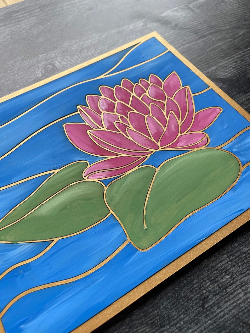Op de afbeelding: Een handgeschilderd houten paneel met een roze waterlelie en groene bladeren op een blauwe achtergrond met gouden accenten. Het paneel heeft een gouden rand.