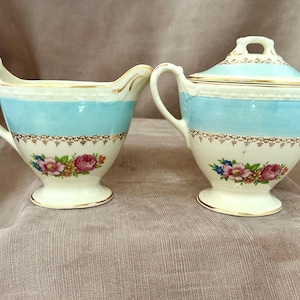 May include: A vintage cream and sugar set. The set features a cream pitcher and a lidded sugar bowl. Both pieces are white with a light blue band, gold trim, and floral accents. The set is likely made of porcelain.