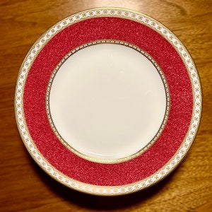 May include: A round, white plate with a red textured border and gold trim. The border features a repeating pattern of gold and white decorative elements. The plate is designed for serving food and is suitable for formal dining.