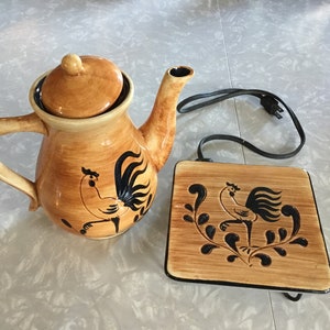 May include: A brown ceramic teapot and matching electric warming plate with a black rooster design. The teapot has a lid and a handle. The warming plate has a cord.
