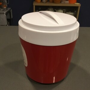May include: A red and white insulated cooler with a white lid and a handle. The cooler is sitting on a gray countertop.