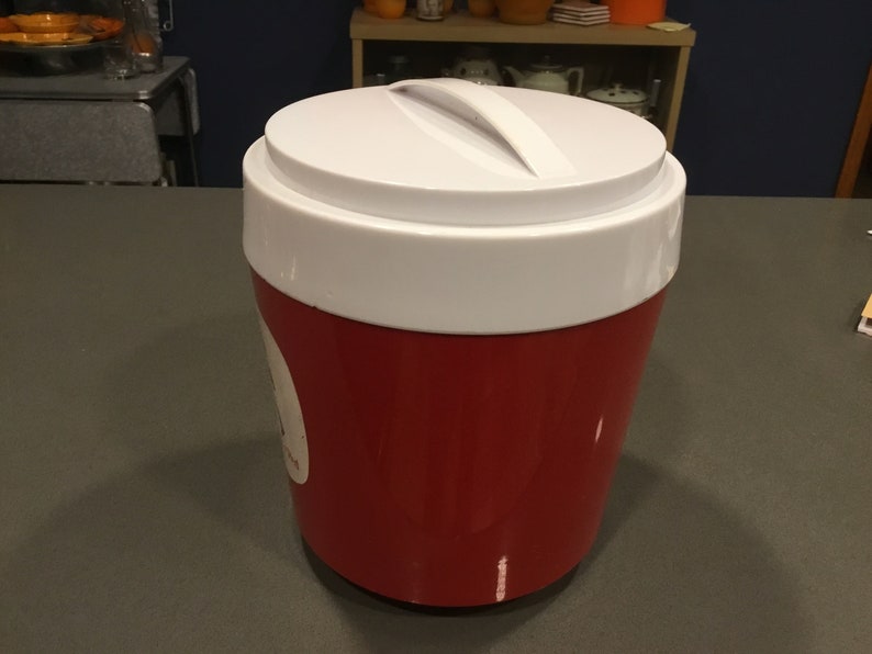 May include: A red and white insulated cooler with a white lid and a handle. The cooler is sitting on a gray countertop.