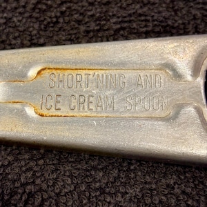 May include: Close-up of a vintage metal Short'ning and Ice Cream Spoon. The spoon has a rectangular handle with the text "SHORT'NING AND ICE CREAM SPOON" embossed on it. The metal has some rust.