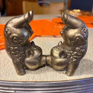 May include: Two silver-toned pig figurines facing each other, connected at the snouts. The pigs have detailed features, including curled tails and ornate designs on their bodies. They are positioned on a patterned surface.