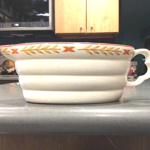 May include: White ceramic serving bowl with a red and gold rim and a wheat design. The bowl has a fluted design and a handle.