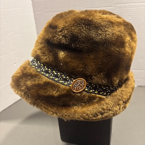 May include: A brown faux fur hat with a short brim. The hat features a black and yellow patterned band and a decorative wooden button. The hat is a vintage style.