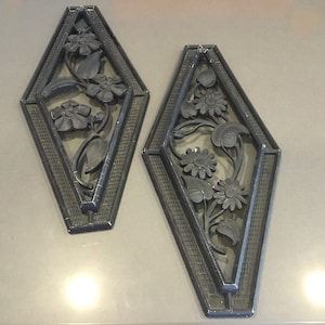 May include: Two matching, dark gray diamond-shaped wall decor pieces. Each features a floral design with detailed leaves and flowers. The borders have a textured pattern, adding depth to the artwork.