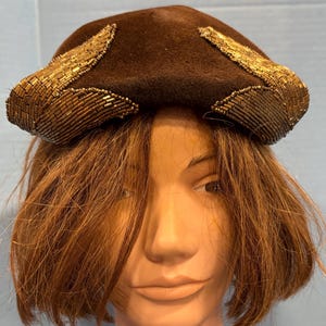 May include: A brown felt hat with gold beaded accents. The hat features a rounded crown and is adorned with beaded embellishments along the brim. The hat is a vintage style and is suitable for special occasions.