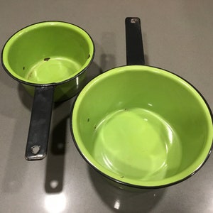 Puede incluir: Dos cacerolas de esmalte verde con asas negras. Las cacerolas están vacías y tienen un borde negro alrededor de la parte superior.