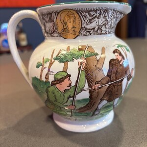 May include: A white ceramic pitcher with a detailed illustration of a hunting scene. The pitcher features a handle, a spout, and a green rim. The artwork depicts figures in green and brown clothing, with trees and an animal. A portrait is on the top.
