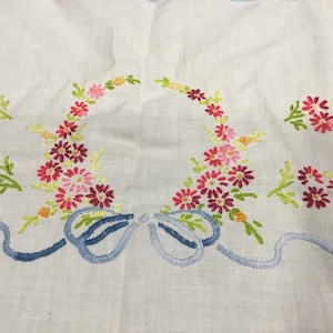 May include: White linen tablecloth with embroidered floral designs in pink, red, and yellow. The flowers are arranged in a wreath shape with a blue ribbon bow in the center.
