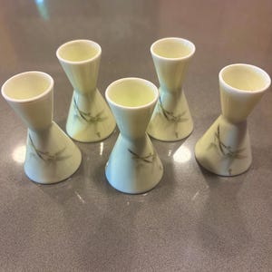May include: Five pale yellow ceramic shot glasses with a flared base and a narrow waist. Each glass features a delicate floral design and a white rim. The glasses are arranged on a gray surface.