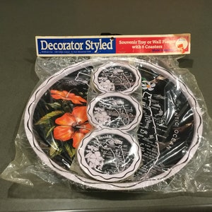 Puede incluir: Un juego de bandeja de recuerdo y seis posavasos, empaquetados en plástico. La bandeja y los posavasos presentan un fondo negro con un mapa de Florida, flores de hibisco naranja y el texto "Florida". La bandeja tiene un borde festoneado blanco.