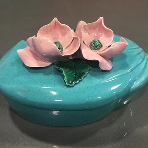 May include: A teal ceramic trinket box with a lid featuring two pink flowers and a green leaf.