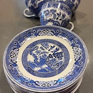 May include: A stack of white and blue Willow pattern plates with matching teacups. The plates and cups feature a detailed landscape design with bridges, trees, and birds. The teacups have handles.