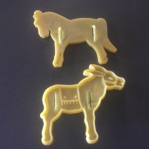 May include: Two yellow plastic cookie cutters, one shaped like a horse and the other shaped like a donkey.
