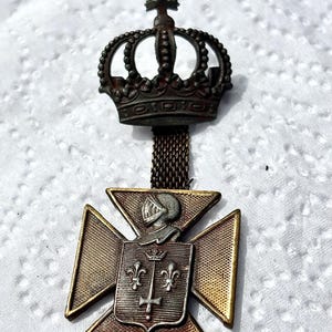 May include: An antique metal pendant featuring a crown and a cross-shaped emblem. The emblem has a knight's helmet, fleur-de-lis, and a cross. The pendant is a mix of bronze and silver tones, with a chain connecting the crown and emblem.