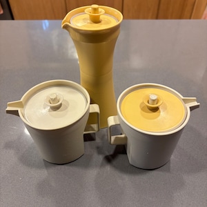 May include: A vintage kitchen set featuring a tall, yellow pitcher with a lid, and two matching cream-colored sugar bowls with lids. The set is made of plastic and has a mid-century modern aesthetic.