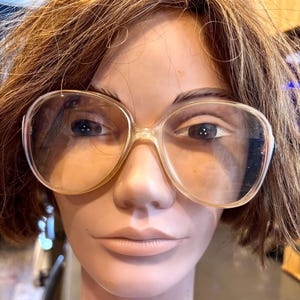 May include: A mannequin head with brown hair and large, clear-lensed eyeglasses. The glasses have a light-colored frame and a retro, oversized design. The mannequin's skin is a pale tone, and the overall aesthetic suggests a vintage or retro style.