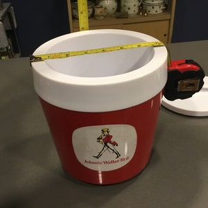 May include: Red and white plastic cooler with a Johnnie Walker Red label featuring a man in a red coat and a walking stick. The cooler has a white lid and a tape measure around the top.