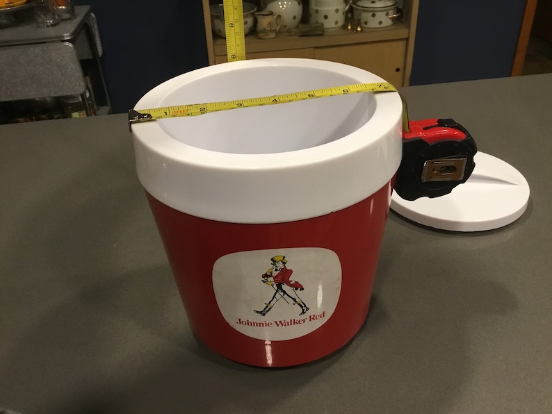 May include: Red and white plastic cooler with a Johnnie Walker Red label featuring a man in a red coat and a walking stick. The cooler has a white lid and a tape measure around the top.