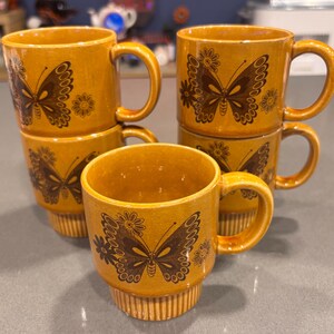 May include: A set of six mustard-yellow ceramic mugs. Each mug features a brown butterfly and floral design. The mugs have a rounded shape with a handle and are stacked in a group. The mugs are suitable for coffee, tea, or other beverages.