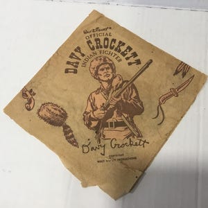 May include: A brown paper napkin with a vintage illustration of Davy Crockett, an "Indian Fighter", holding a rifle. The text "Walt Disney's Official Davy Crockett Indian Fighter" is printed in brown lettering. The napkin has a brown raccoon tail and a brown knife illustration.