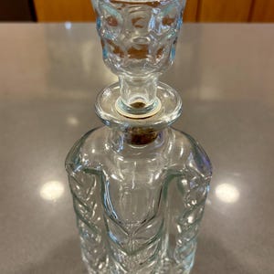 May include: A clear glass decanter with a decorative stopper. The bottle has a leaf-like design on the sides and a round base. The stopper has a floral pattern. The decanter is empty and sits on a gray surface.