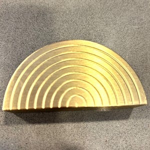 May include: A brass-colored, semi-circular object with concentric, curved grooves. The design resembles a stylized rainbow or arch. The object is likely a decorative element or hardware component.