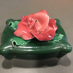 May include: A green ceramic trinket box with a pink rose on top. The rose is made of ceramic and has a glossy finish.
