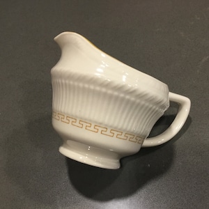 May include: White ceramic creamer with a gold Greek key design around the top edge. The creamer has a fluted design and a handle.