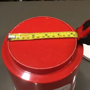 May include: Red plastic container with a lid. The lid has a yellow measuring tape around it, measuring 10 inches in diameter.