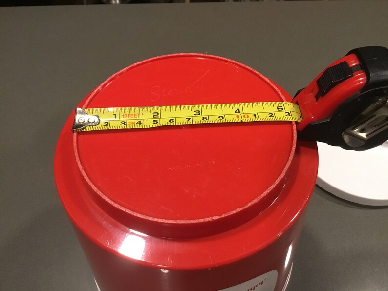 May include: Red plastic container with a lid. The lid has a yellow measuring tape around it, measuring 10 inches in diameter.