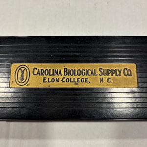 May include: A black, grooved rectangular object with a gold-colored rectangular plate. The plate reads "CAROLINA BIOLOGICAL SUPPLY CO. ELON COLLEGE, N.C." in black text. The background is a light, off-white color.