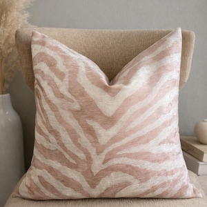 May include: A decorative square pillow with a zebra stripe pattern in shades of blush pink and cream. The pillow is placed on a beige chair, creating a soft and inviting aesthetic. The pillow's design adds a touch of modern flair to the room.