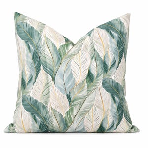 May include: A square decorative pillow with a botanical leaf pattern. The pillow features a repeating design of large, stylized leaves in shades of green, blue, and cream, with gold accents. The leaves overlap, creating a layered effect against a white background.