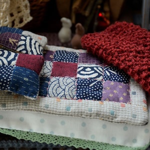 Pode incluir: Uma cama pequena com uma colcha de retalhos e um cobertor de malha vermelho. A colcha é feita de tecido azul e branco com uma variedade de padrões. A cama é feita de madeira e tem um lençol branco.