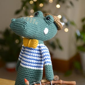 Könnte beinhalten: Gehäkelte Alligator-Puppe mit grünem Körper, blau-weiß gestreiftem Oberteil, gelber Fliege und braunem Hut. Ein handgefertigtes, charmantes Spielzeug.