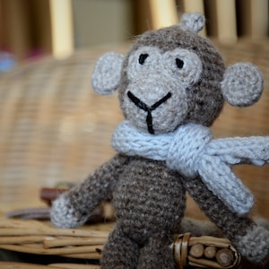 May include: A crocheted monkey toy wearing a light blue scarf. The monkey has brown fur and black eyes.