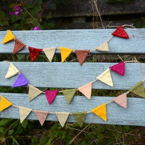 May include: Colorful felt triangle bunting in shades of yellow, red, brown, beige, purple, pink, and green. The pennants are strung on a natural twine and displayed on a weathered blue bench. A decorative item for parties and celebrations.