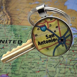 May include: A silver keychain with a round pendant featuring a map of San Antonio, Texas. The map shows major roads and the text "San Antonio". A gold key rests beside the pendant, all set against a map background.
