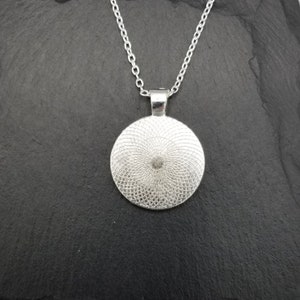 May include: Silver pendant necklace with a round, silver, patterned charm. The charm has a circular design with a raised center.