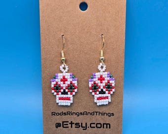 May include: A pair of beaded sugar skull earrings with white, red, green, and purple beads. The earrings are on a brown card with the text "RodsRingsAndThings @Etsy.com"