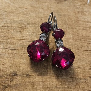 May include: A pair of dangle earrings featuring large, faceted, heart-shaped, deep magenta crystals. Each earring has a smaller magenta stone and a clear crystal accent. The earrings are set in a silver-toned metal.